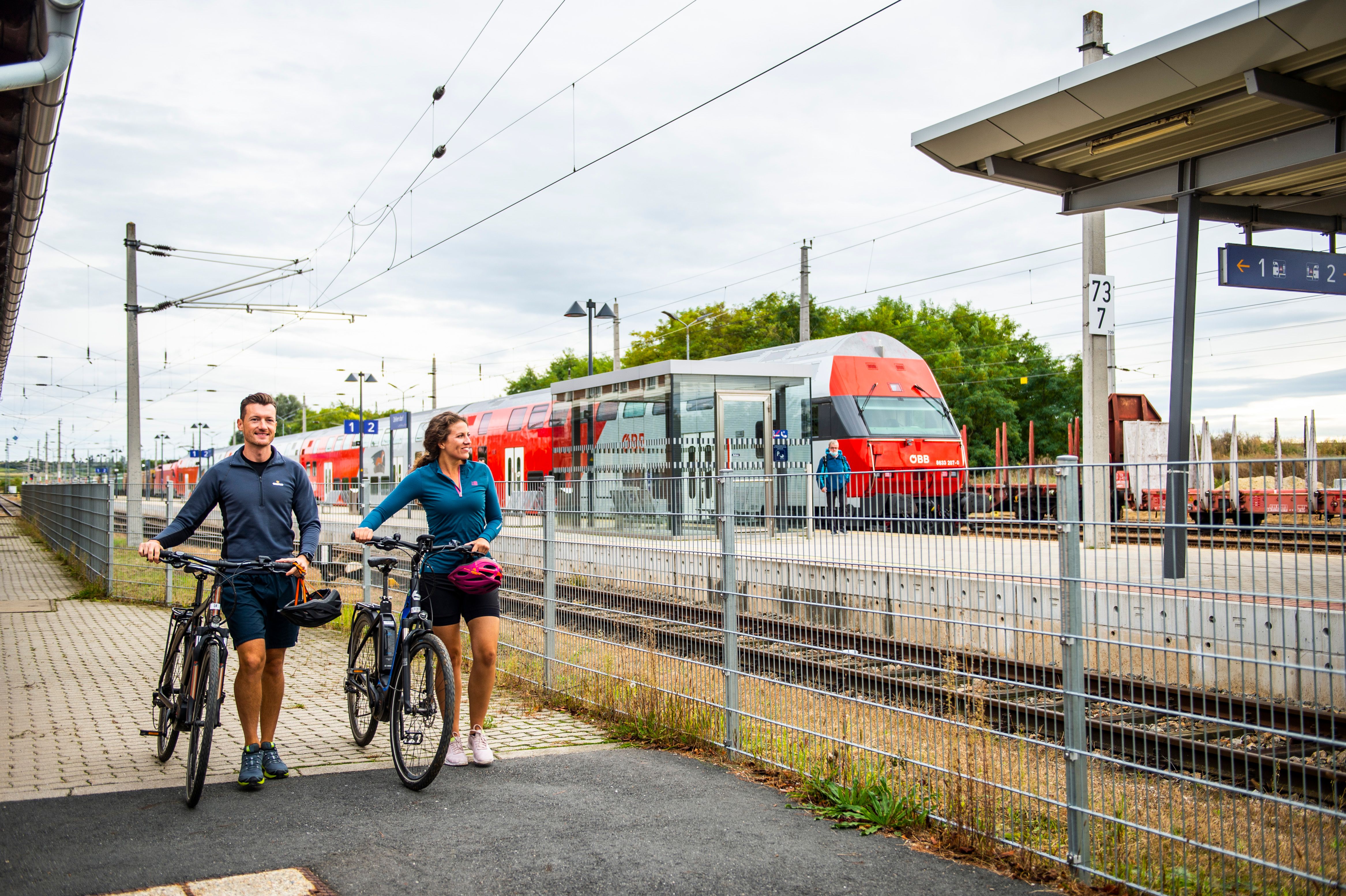 Zwei Personen mit Fahrrädern am Bahnhof Retz, ein ÖBB-Zug im Hintergrund.