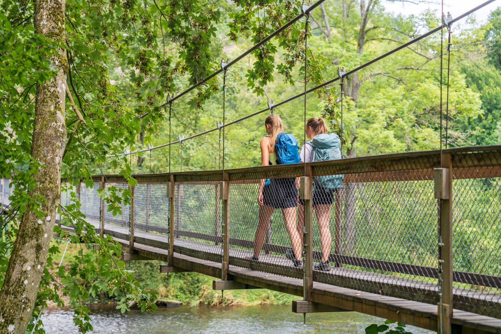 Zwei Wanderinnen mit Rucksäcken gehen über eine Hängebrücke im grünen Wald über einem Fluss.
