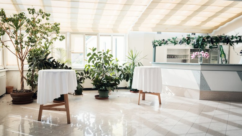 Heller Raum mit Pflanzen, zwei Stehtischen mit weißen Tischdecken und einer Bar im Hintergrund.