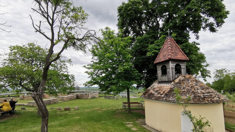 Kleine Kapelle mit rotem Ziegeldach in einer gr&uuml;nen Landschaft, umgeben von B&auml;umen und B&auml;nken.