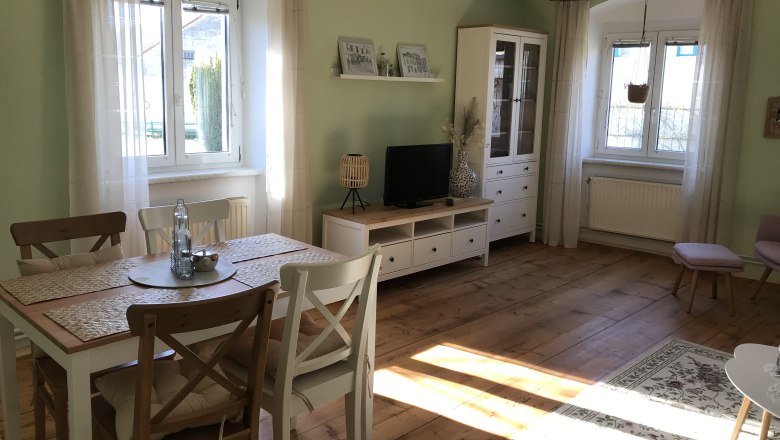 Heller Raum mit Esstisch, St&uuml;hlen, TV-Schrank und gro&szlig;en Fenstern.