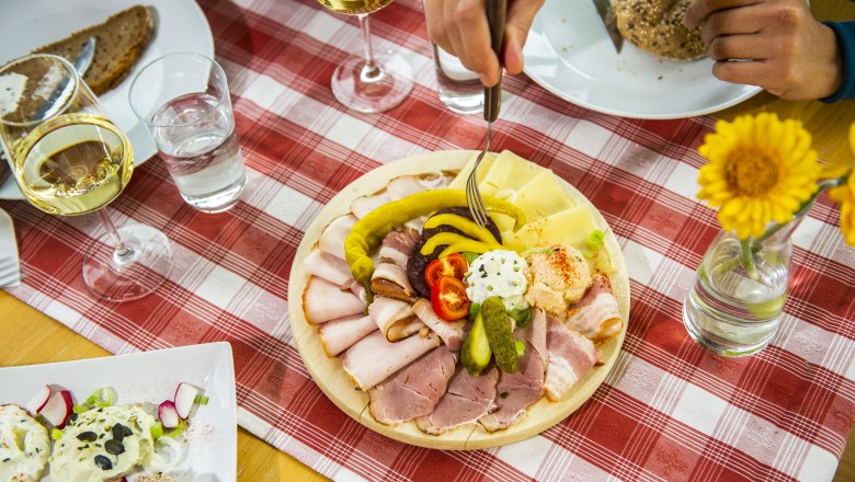 Ein gedeckter Tisch mit einer Platte voller Aufschnitt, Käse und Gemüse, dazu Weißwein und Brot auf einer rot-weiß karierten Tischdecke.