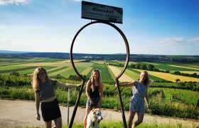 Drei Frauen und ein Hund posieren vor einem gro&szlig;en Schl&uuml;ssellochrahmen mit Blick auf eine l&auml;ndliche Landschaft.