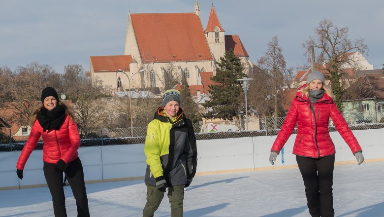 Three people skating on an ice rink with a church in the background.