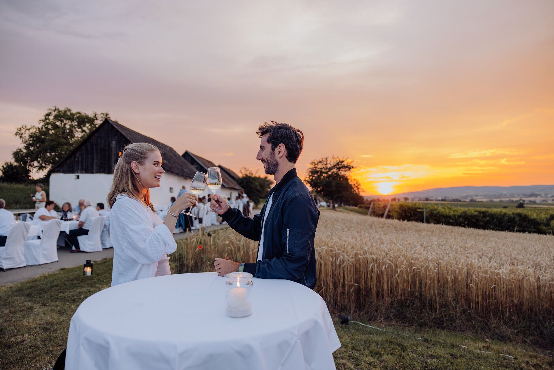 Bei Sonnenuntergang stoßen zwei Freunde mit Gläsern an und genießen den Moment inmitten der goldenen Felder. Die sanften Hügel und die malerische Landschaft schaffen eine einladende Atmosphäre, die zum Verweilen einlädt. Kulinarische Köstlichkeiten und erlesene Weine versprechen ein unvergessliches Erlebnis.