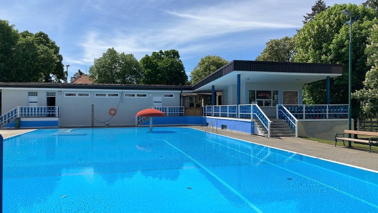 Ein Freibad mit klarem Wasser, umgeben von B&auml;umen und einem Geb&auml;ude mit blauen Gel&auml;ndern.