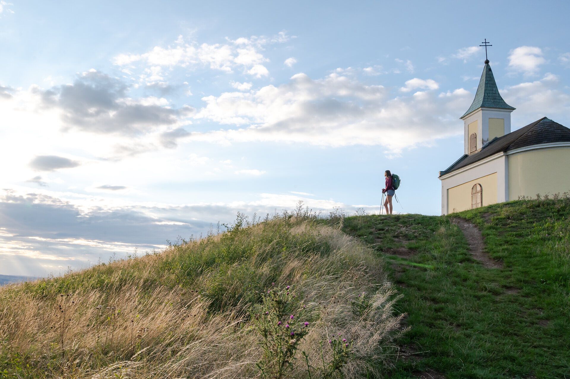 Eine Wanderin mit Rucksack und Wanderstöcken steht am Michelberg neben der Kapelle und genießt den Ausblick.