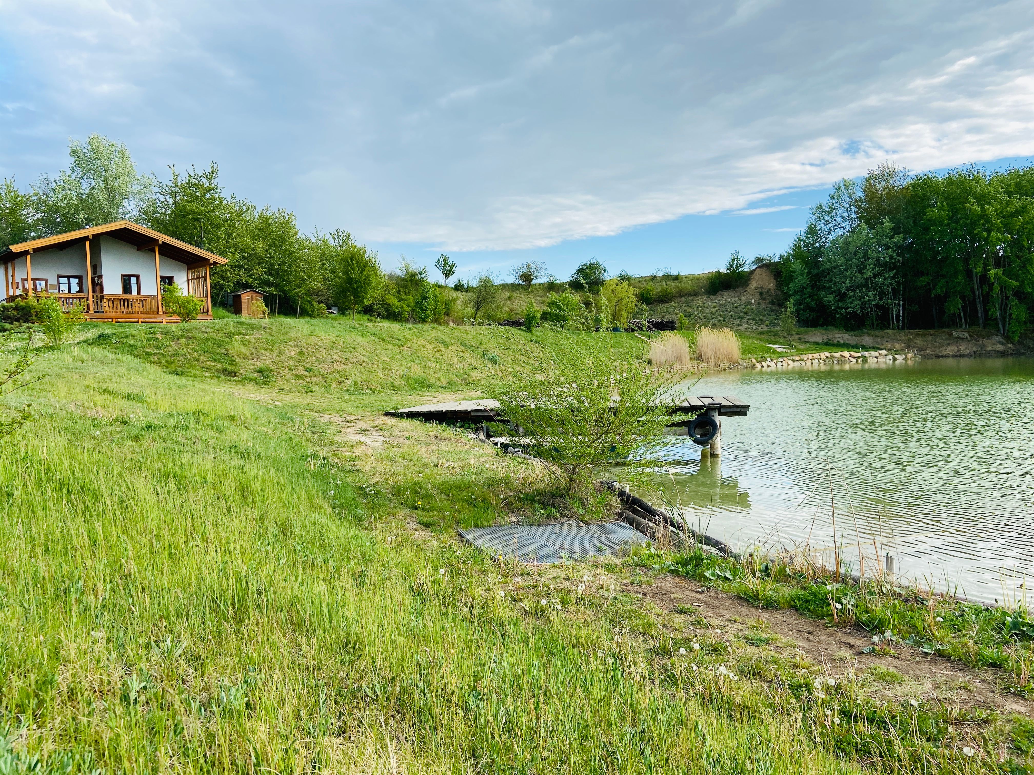 Ein kleines Haus am Ufer eines Teiches mit grüner Wiese und Bäumen im Hintergrund.