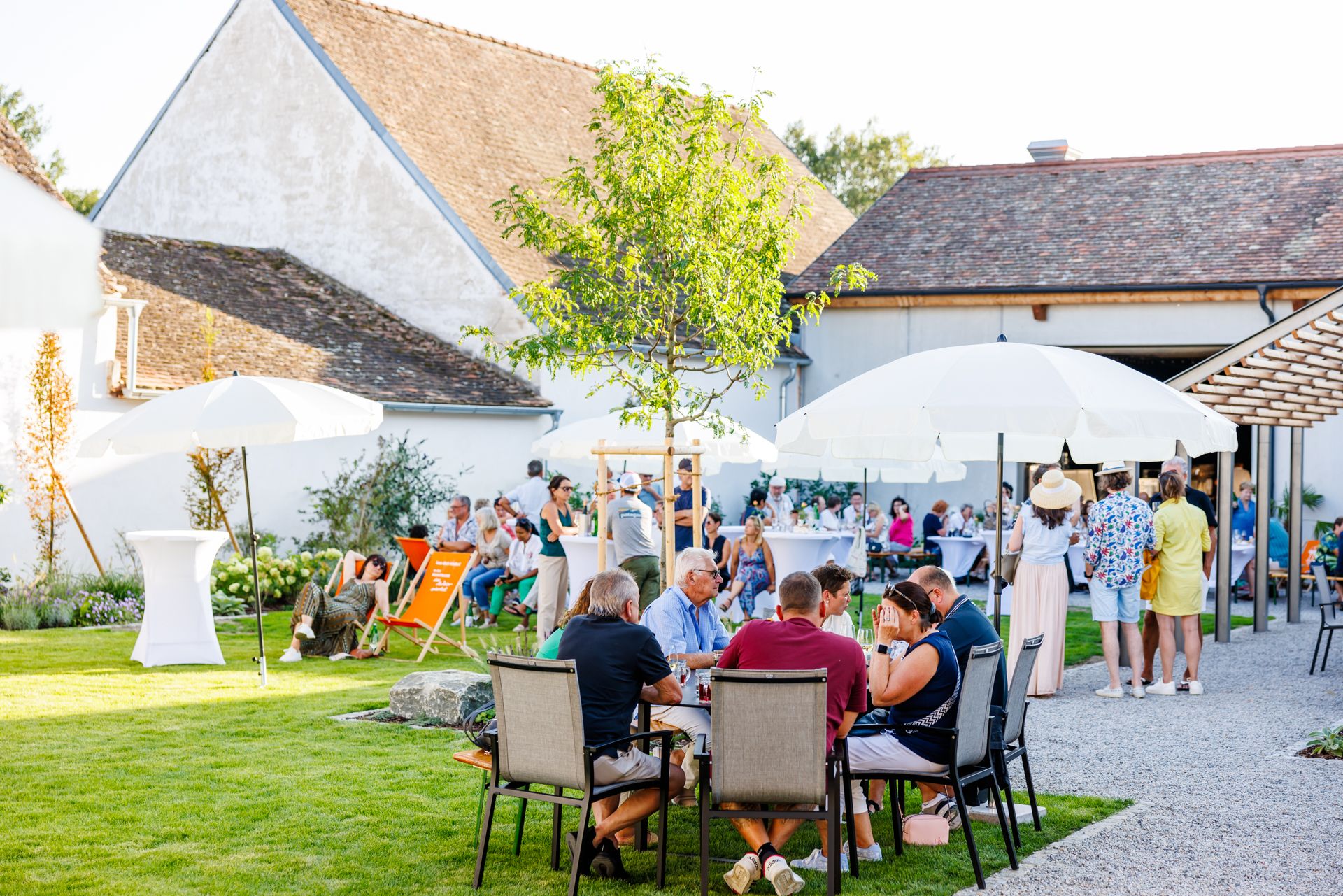 Menschen genießen eine Veranstaltung im Freien mit Tischen, Stühlen und Sonnenschirmen auf einer Wiese vor einem Gebäude.