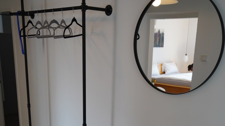 Innenansicht eines Zimmers mit Kleiderst&auml;nder, rundem Spiegel und kleinem Tisch mit Pflanze und Vase.