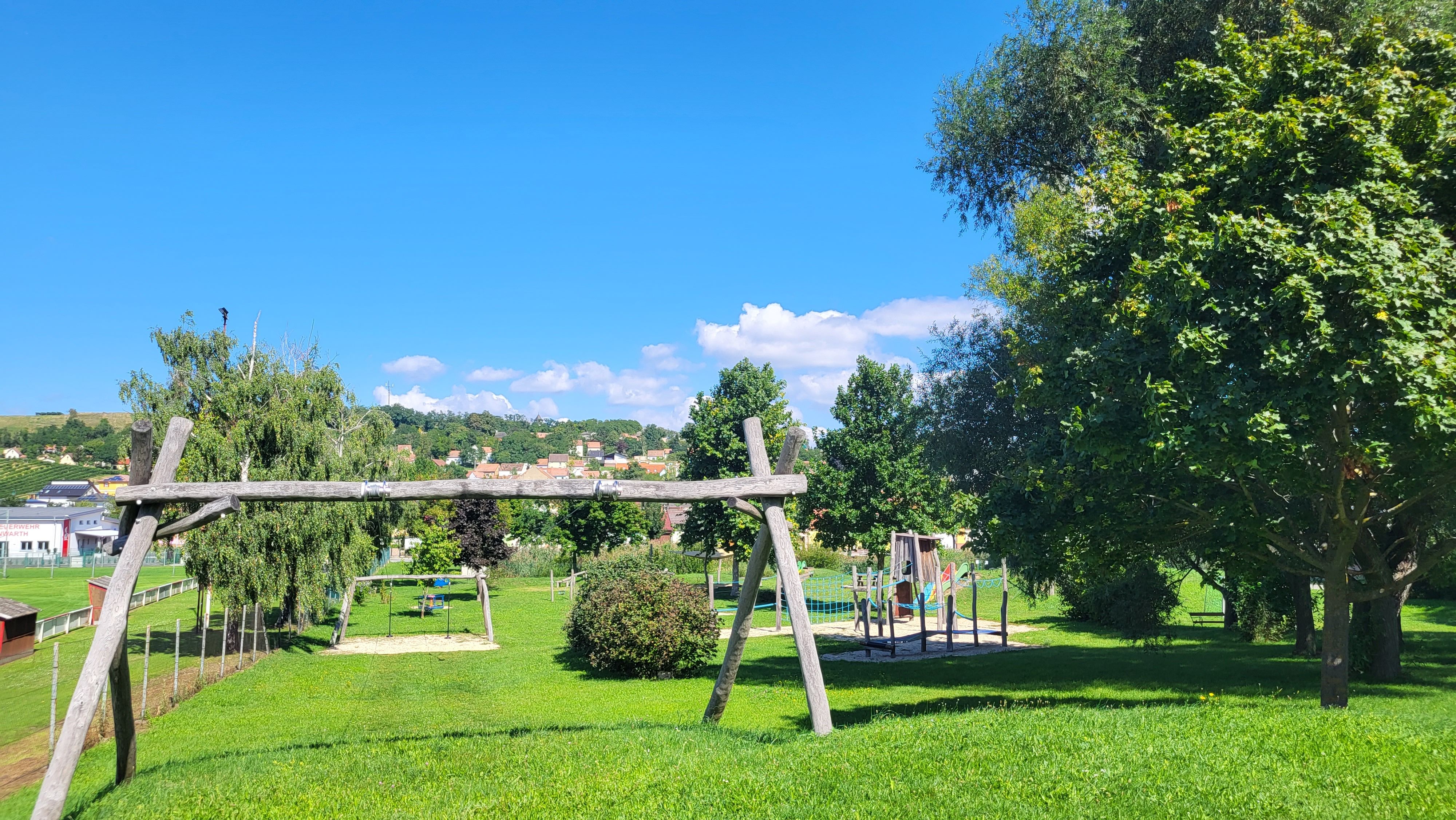 Ein Spielplatz mit Schaukeln und Klettergerüst auf einer grünen Wiese, umgeben von Bäumen und einem klaren blauen Himmel.