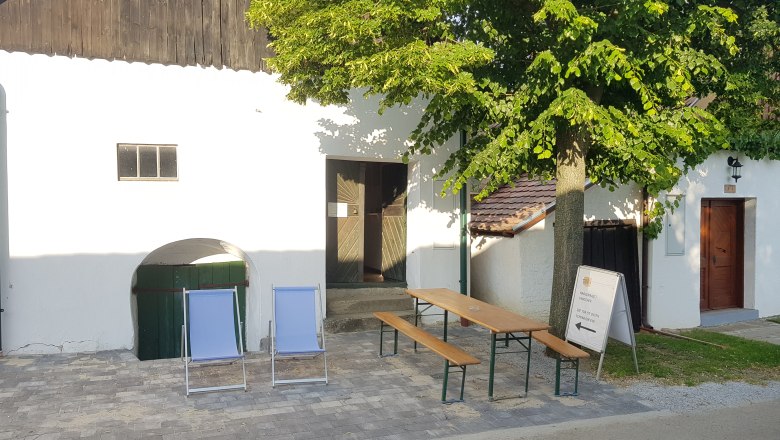 Zwei Liegestühle und ein Tisch mit Bänken vor einem weißen Gebäude mit Baum.