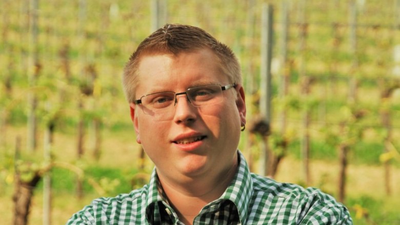 Ein Mann mit Brille und kariertem Hemd steht mit verschränkten Armen vor einem Weinberg.