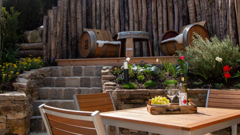 A cozy garden area with wooden table and chairs, wine bottles and glasses. A wooden wall in the background.