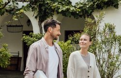 Ein Paar schlendert entspannt durch die malerische Umgebung eines Winzerhofs, während sie einen Weinkarton in der Hand halten. Die sanften Hügel und üppigen Weinreben schaffen eine einladende Atmosphäre, die zum Genießen und Verweilen einlädt.