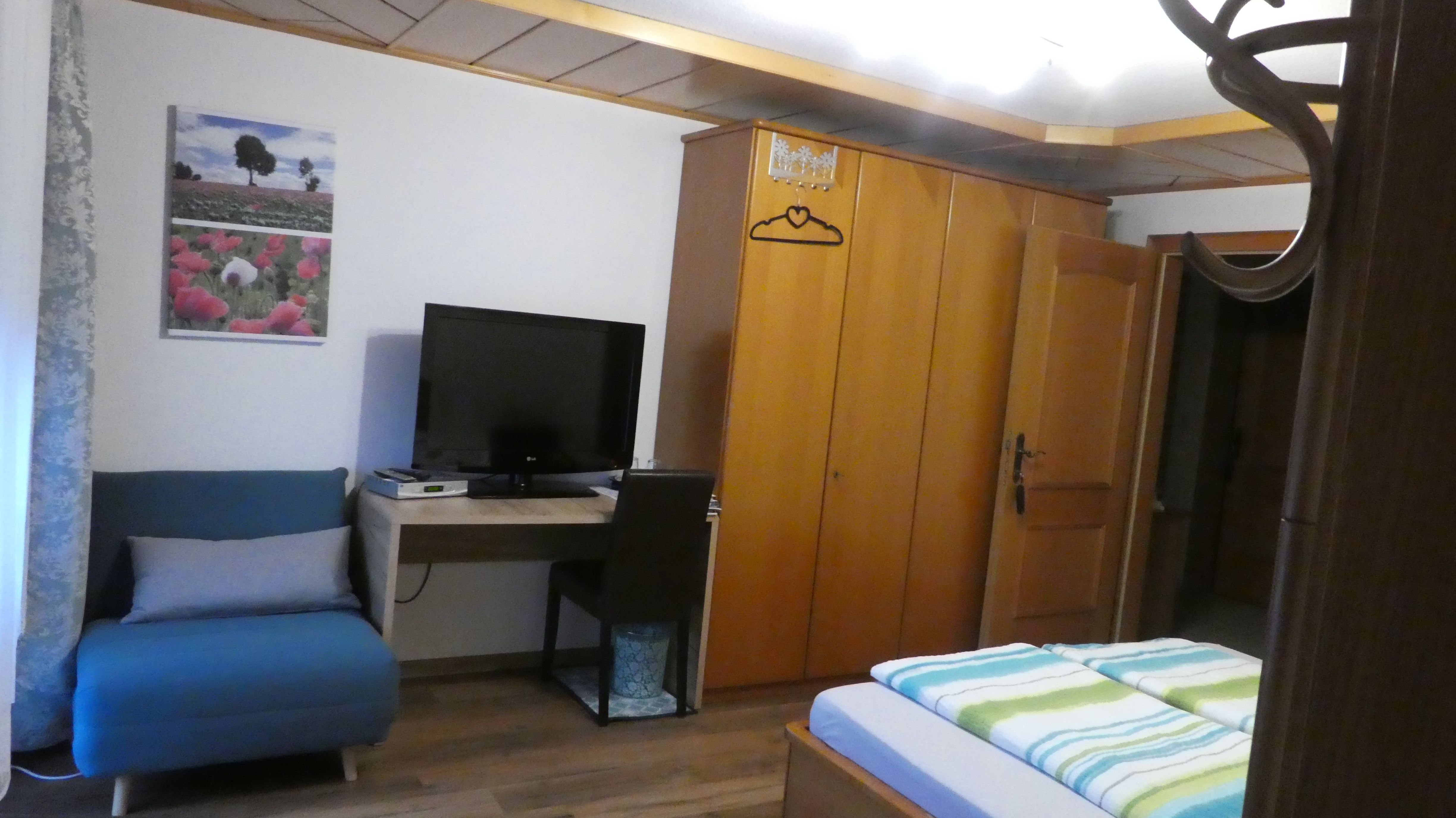 Ein gemütliches Privatzimmer mit einem blauen Sessel, einem Fernseher auf einem Schreibtisch, einem Kleiderschrank und einem Bett mit gestreifter Bettwäsche.