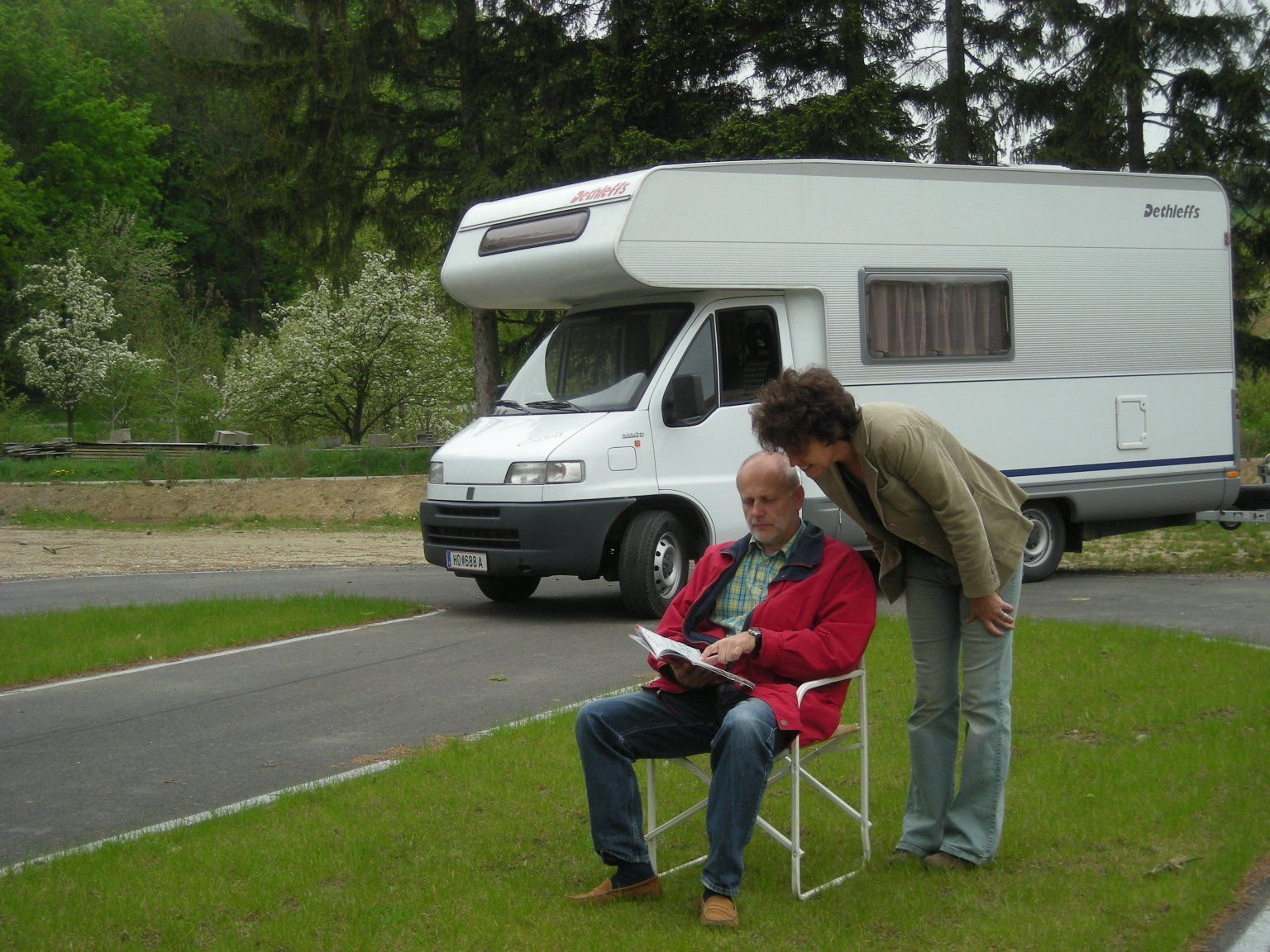 Ein Mann sitzt auf einem Campingstuhl vor einem Wohnmobil, während eine Frau über seine Schulter schaut. Im Hintergrund sind Bäume und Wiese zu sehen.
