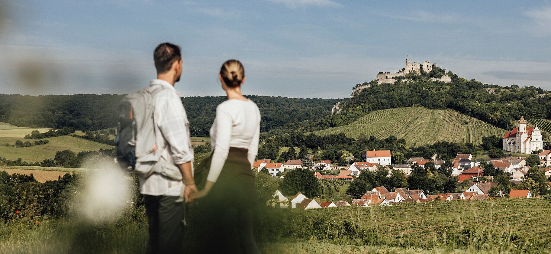 Ein romantisches Paar genießt die atemberaubende Aussicht auf die sanften Weinberge, die sich bis zur majestätischen Burgruine Falkenstein erstrecken. Die warmen Sonnenstrahlen und die frische Luft des Weinfrühlings schaffen eine einladende Atmosphäre für unvergessliche Erlebnisse in der Natur.
