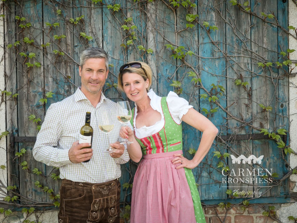 Ein Mann und eine Frau in traditioneller Kleidung halten Weinflaschen und Gläser vor einer bewachsenen Holzwand.