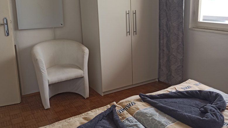 Ein Gästezimmer mit zwei Betten, einem weißen Schrank, einem Sessel und einem Fenster mit Vorhang.