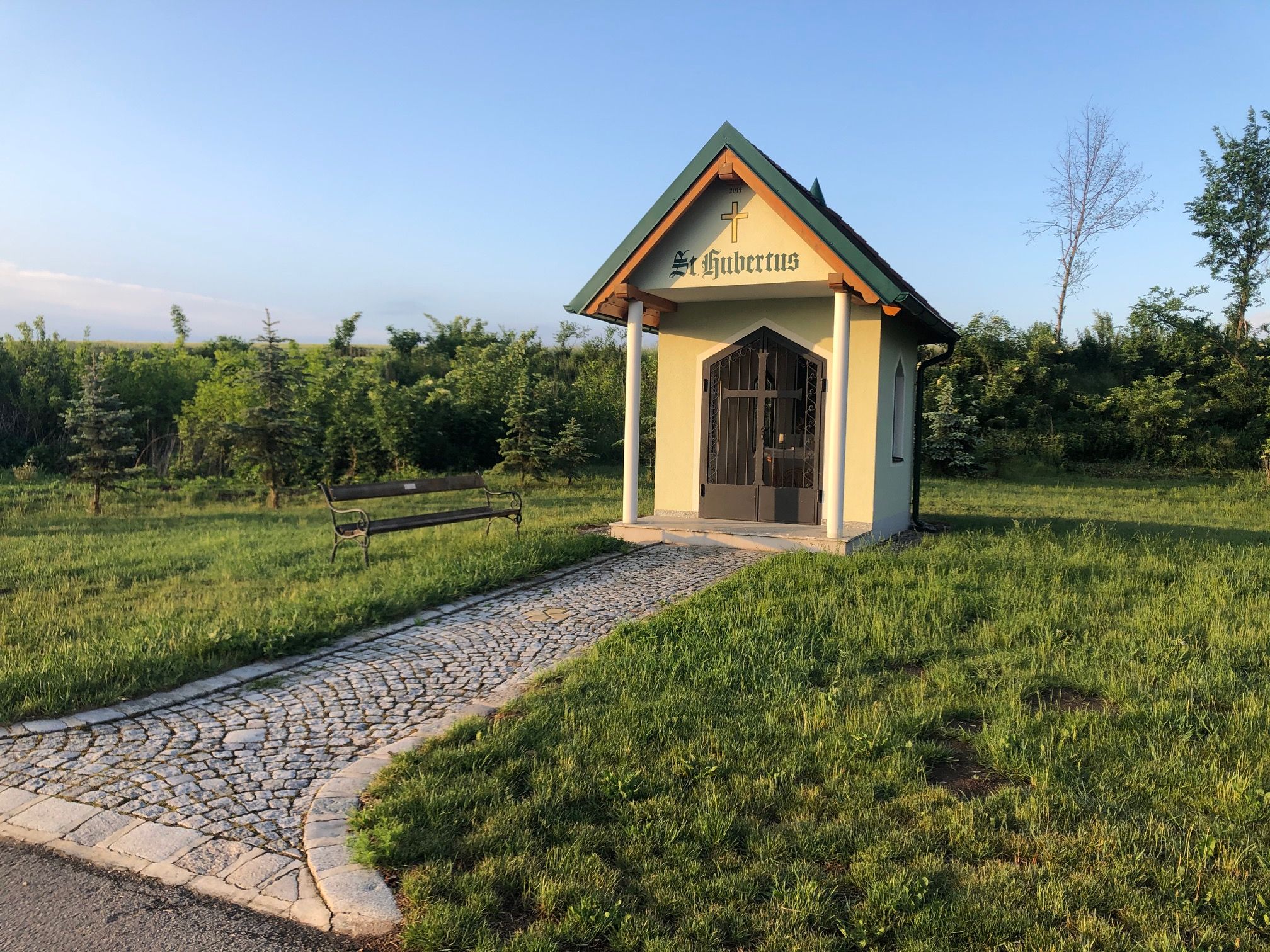 Kleine Kapelle mit Kreuz und Schriftzug "St. Hubertus" auf einer Wiese, mit gepflastertem Weg und Bank.