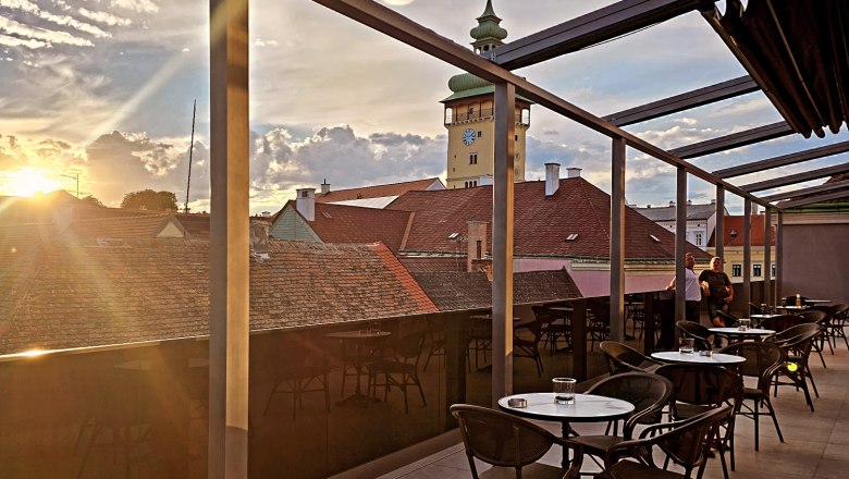 Rooftop-Terrasse mit Tischen und Stühlen, Blick auf den Rathausturm und Sonnenuntergang.