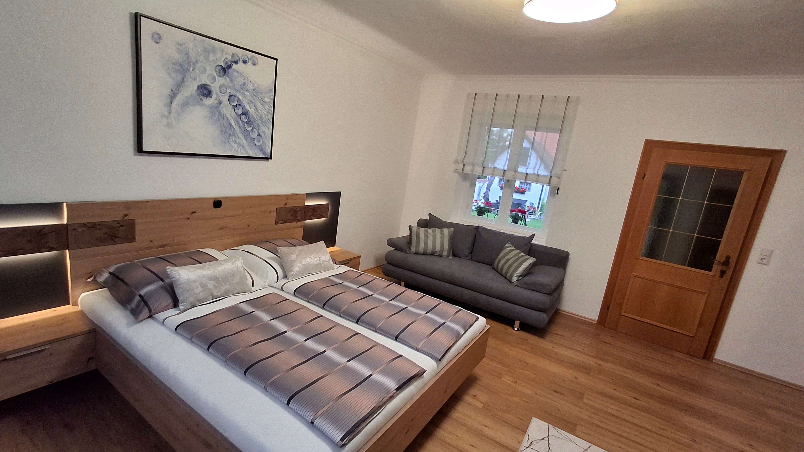 Modernes Schlafzimmer mit Doppelbett, grauem Sofa, Holzfußboden und abstraktem Kunstwerk an der Wand.