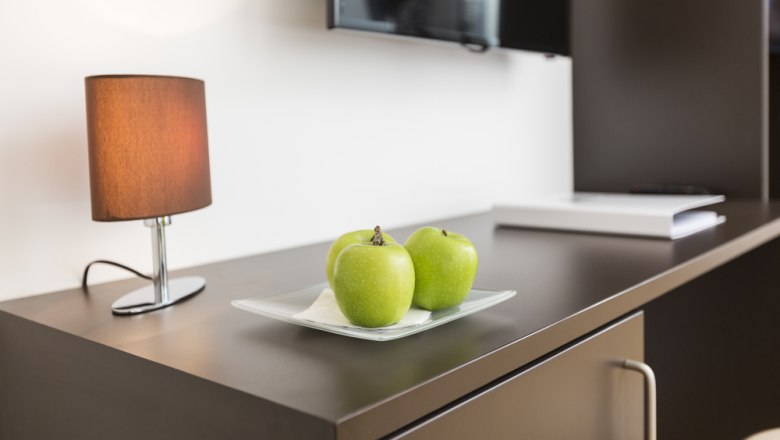 Ein Tisch mit drei grünen Äpfeln auf einem Teller, einer Lampe und einem Fernseher im Hintergrund.