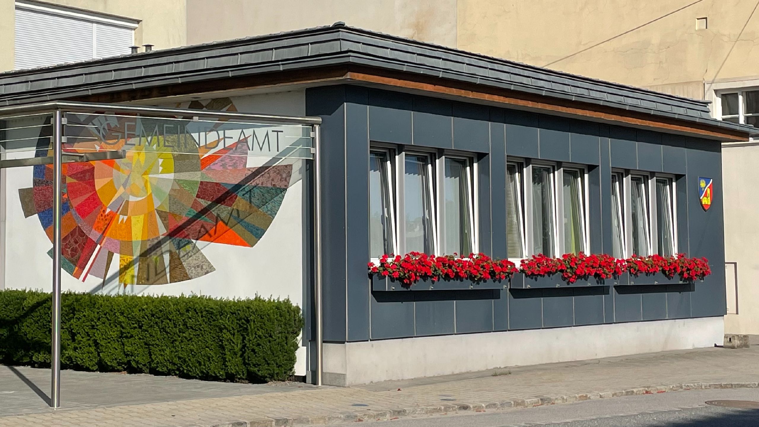 Gemeindeamt mit moderner Fassade, buntem Glasbild, roten Blumenkästen und Wappen an der Wand.