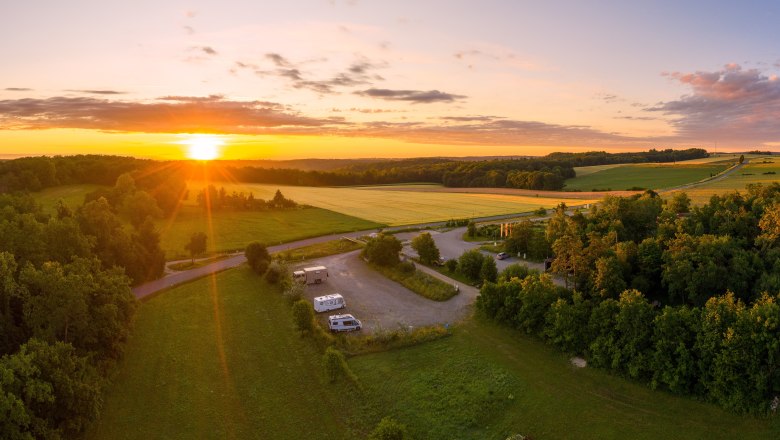 Aerial view of a motorhome site at sunset, surrounded by fields and woods.
