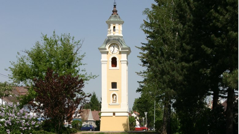 Gelber Kirchturm mit Uhr in Hohenau, umgeben von Bäumen.