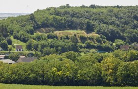 Landschaft mit Hügel, Bäumen und Häusern im Vordergrund.