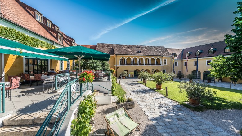 Ein sonniger Innenhof mit gelben Gebäuden, Terrassenbereich und Liegestühlen.