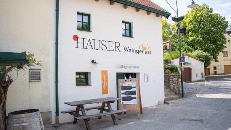 Außenansicht eines Gebäudes mit der Aufschrift 'Hauser Chalet Weingenuss'. Vor dem Gebäude steht ein Holztisch und ein Weinfass.