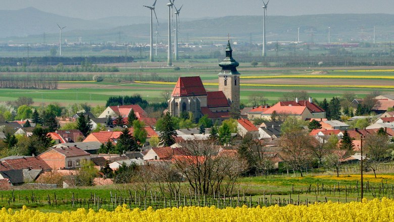 Blick auf Pillichsdorf mit Kirche, umgeben von Feldern und Windrädern im Hintergrund.