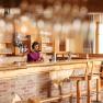 A woman stands behind a bar in a rustic restaurant and pours wine.