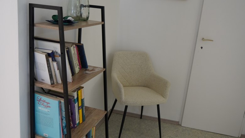 Ein B&uuml;cherregal mit B&uuml;chern und Dekoration, daneben ein beiger Stuhl und eine T&uuml;r in einem Raum.