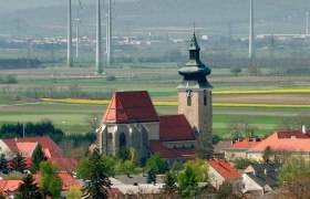 Kirche in Pillichsdorf mit umliegenden Häusern und Windrädern im Hintergrund.