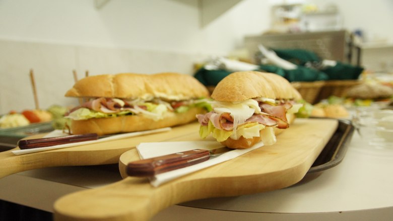 Zwei belegte Baguettes auf Holzbrett mit Messer, im Hintergrund weitere Speisen.