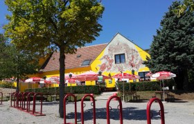 Ein gelbes Gebäude mit Wandmalerei, umgeben von Bäumen und roten Sonnenschirmen.