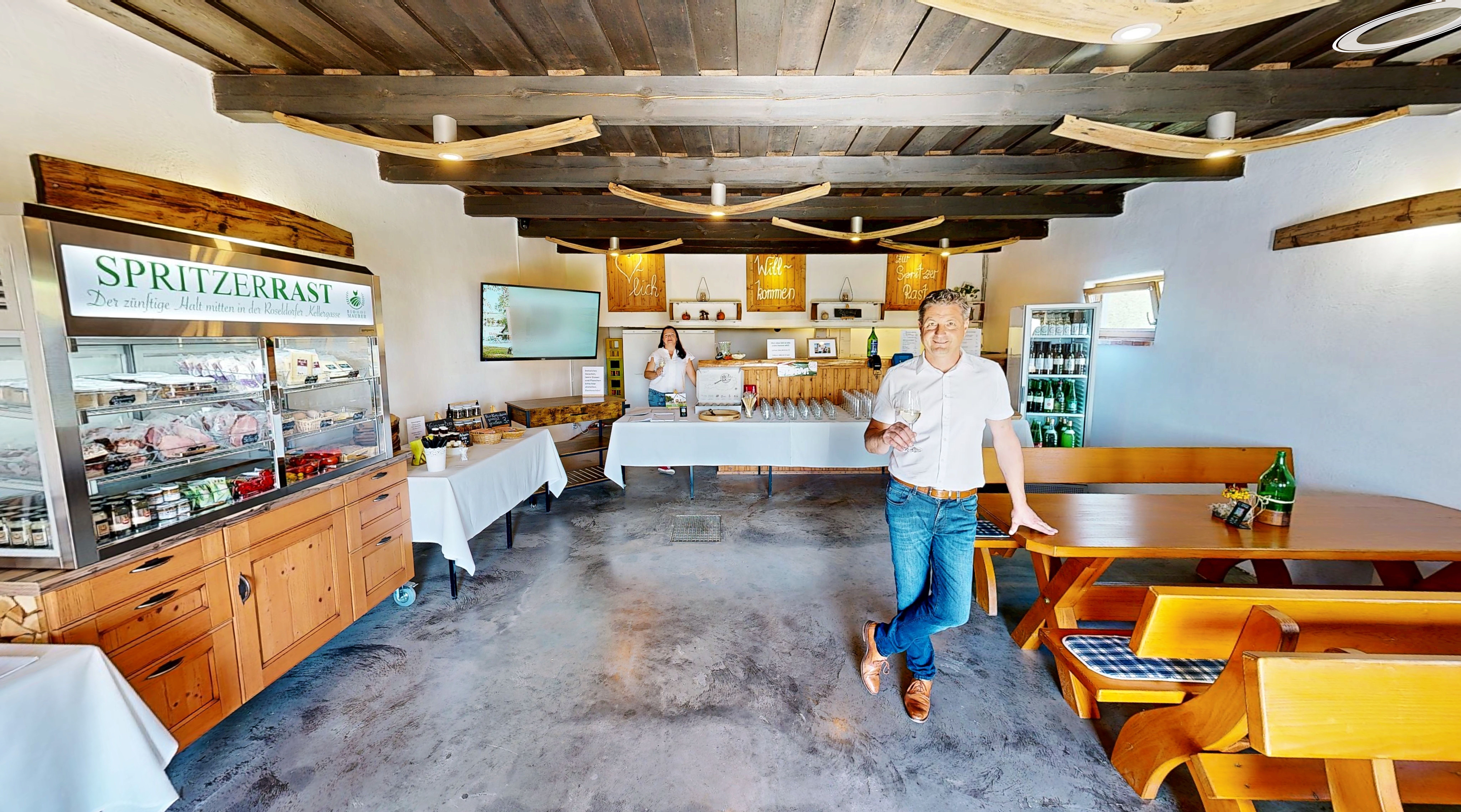 Ein Mann steht in einem rustikalen Raum mit Holzmöbeln und einem Kühlschrank voller Getränke. Ein Schild mit der Aufschrift 'Spritzerrast' ist sichtbar.