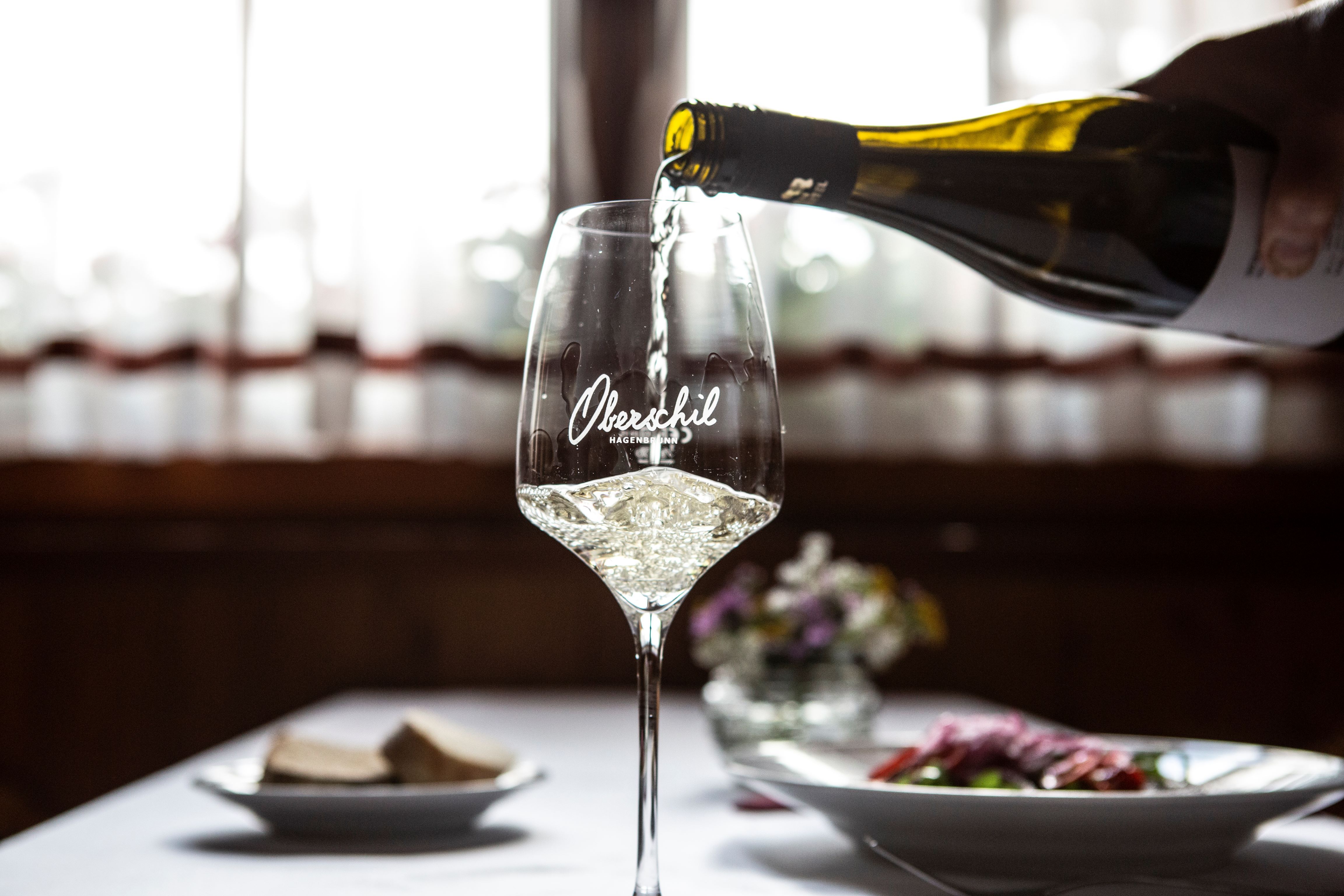 Ein Glas Weißwein wird eingeschenkt, im Hintergrund ein gedeckter Tisch mit Blumen.