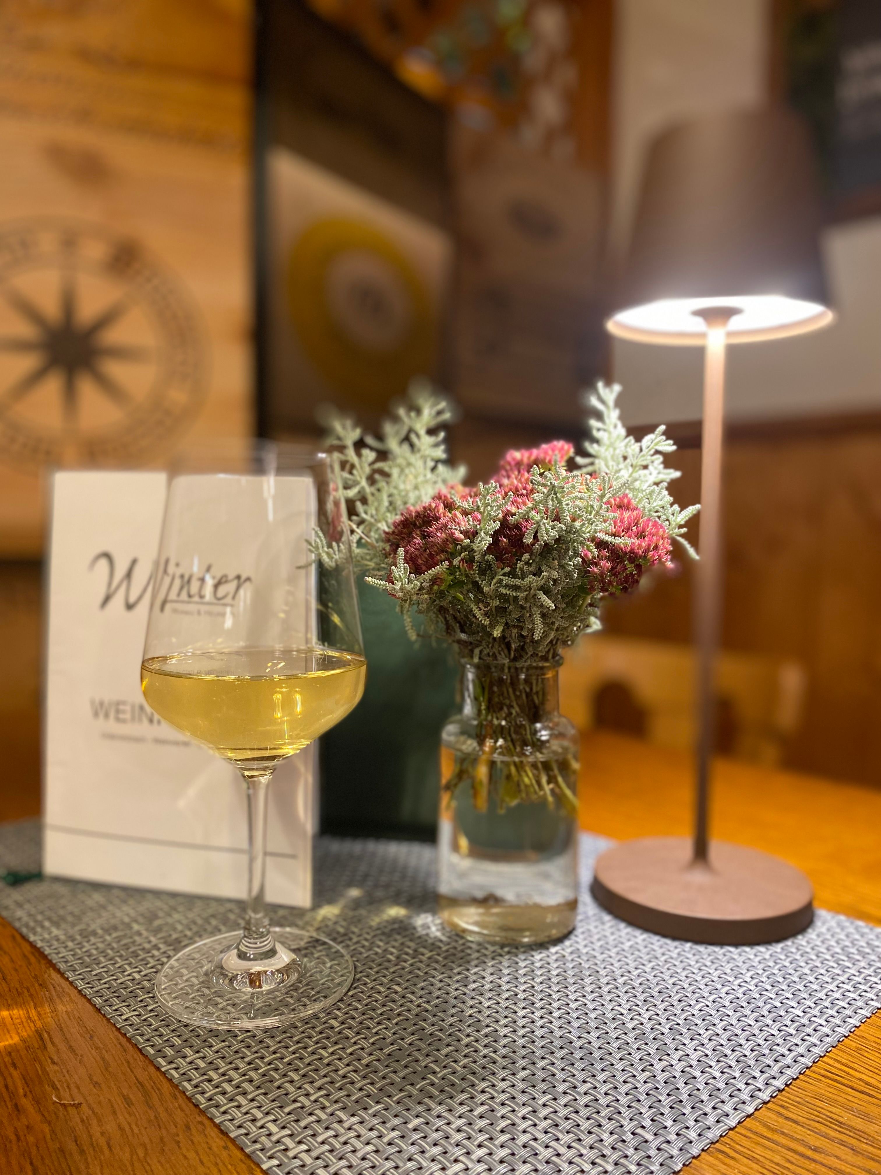 Ein Weinglas mit Weißwein steht auf einem Tisch neben einer Vase mit Blumen und einer Tischlampe.