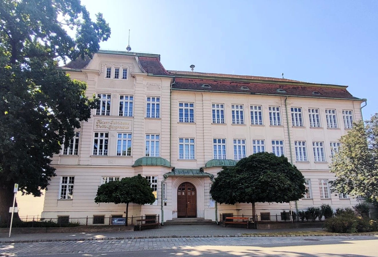 Pohled zepředu na školu „Kaiser Jubiläumsschule“ v Haugsdorfu se stromy v popředí.