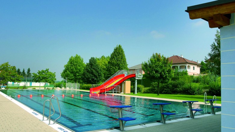 Ein Freibad mit einem Schwimmbecken, einer roten Rutsche und Startblöcken. Im Hintergrund sind Bäume und ein Wohnhaus zu sehen.