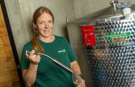 Eine Frau in einem gr&uuml;nen T-Shirt steht neben einem gro&szlig;en Edelstahltank und h&auml;lt ein Glas mit einer Fl&uuml;ssigkeit.