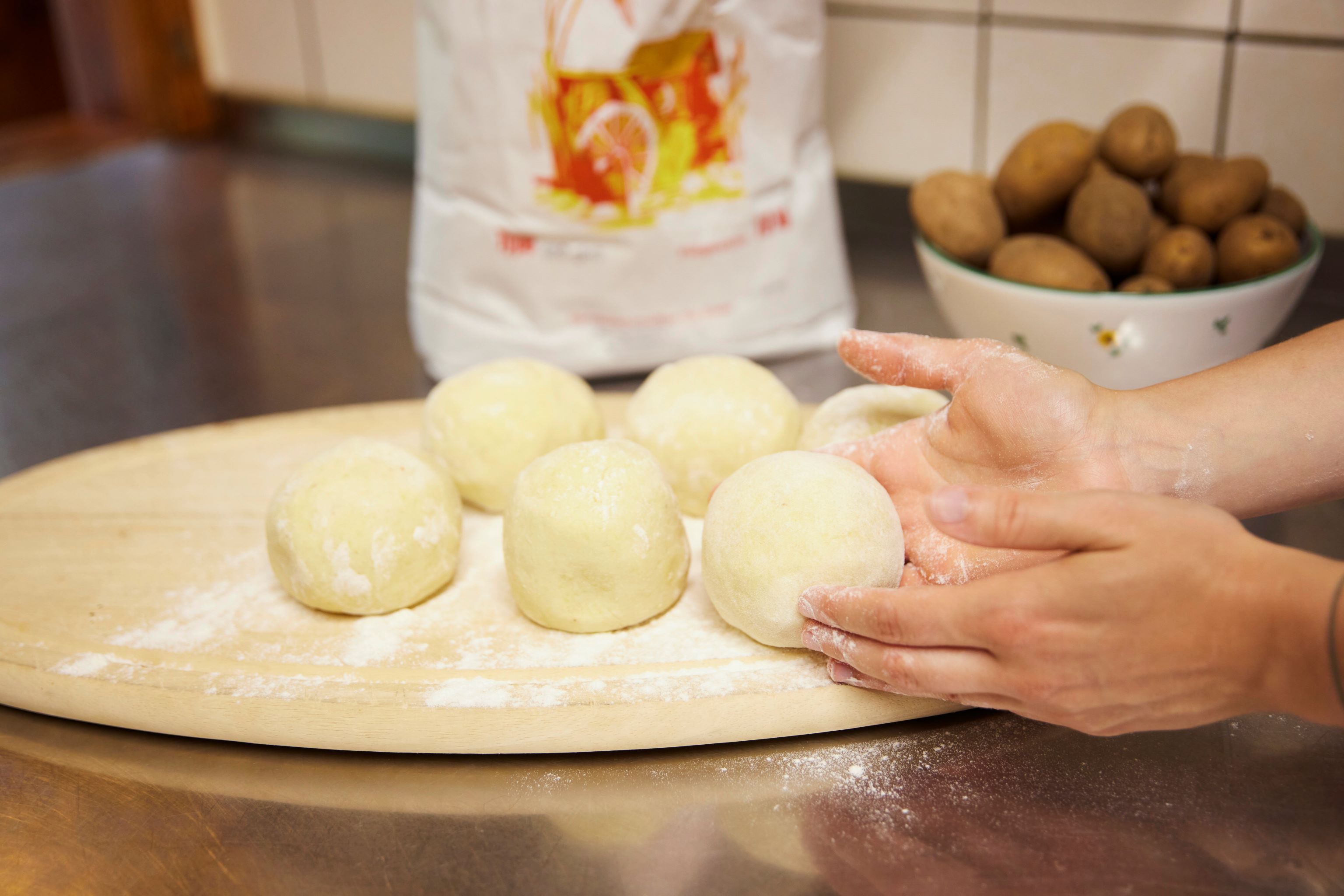 Hände formen Knödelteig auf einem bemehlten Holzbrett, im Hintergrund eine Schüssel mit Kartoffeln.