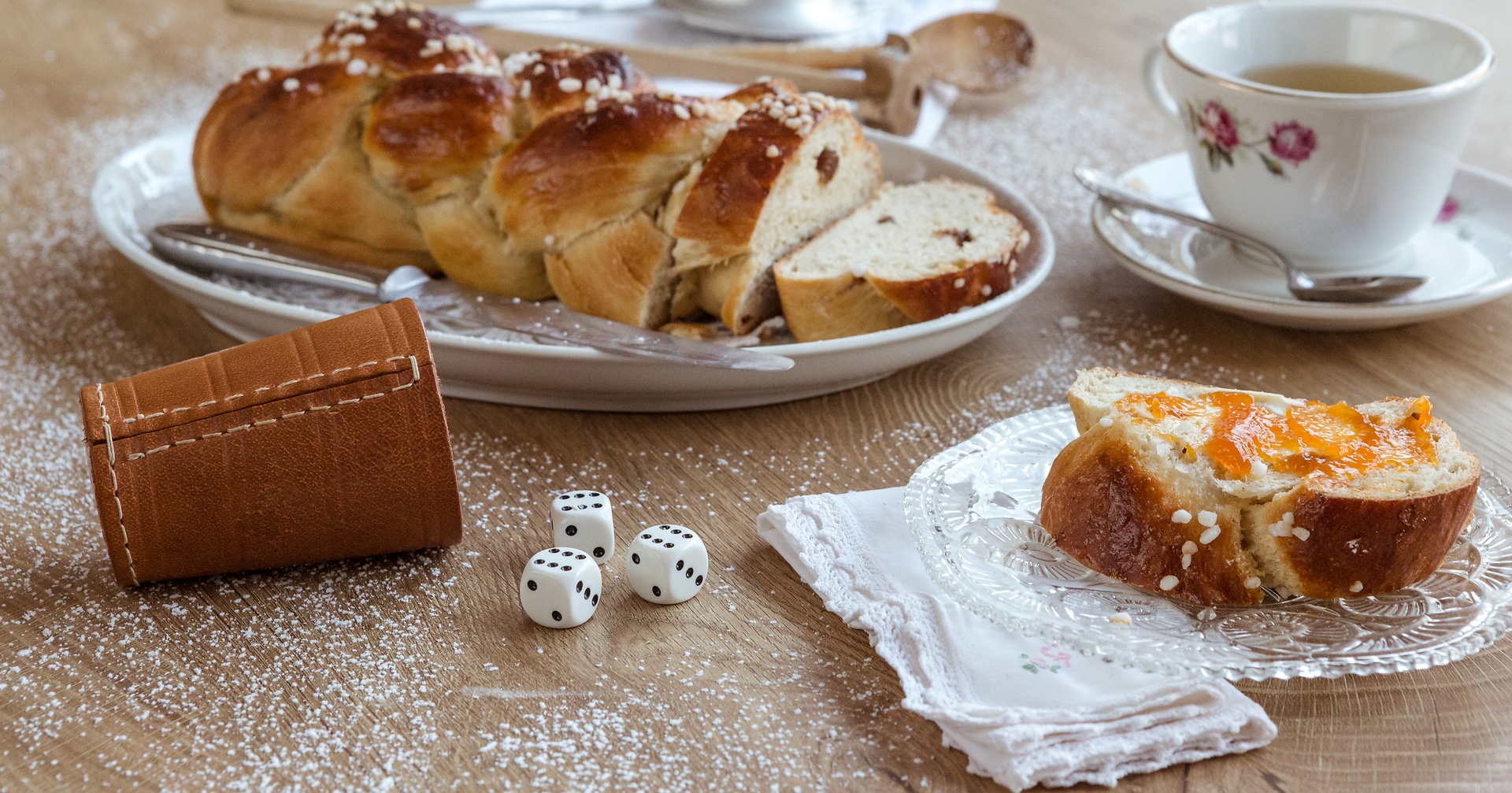 Ein frisch gebackener Striezel, goldbraun und mit Zuckerguss verziert, lädt zum Genießen ein. Dazu eine Tasse aromatischer Tee, die die Gemütlichkeit perfekt macht. Die rustikale Holzoberfläche und die kleinen Details wie die Würfel und das Besteck schaffen eine einladende Atmosphäre.