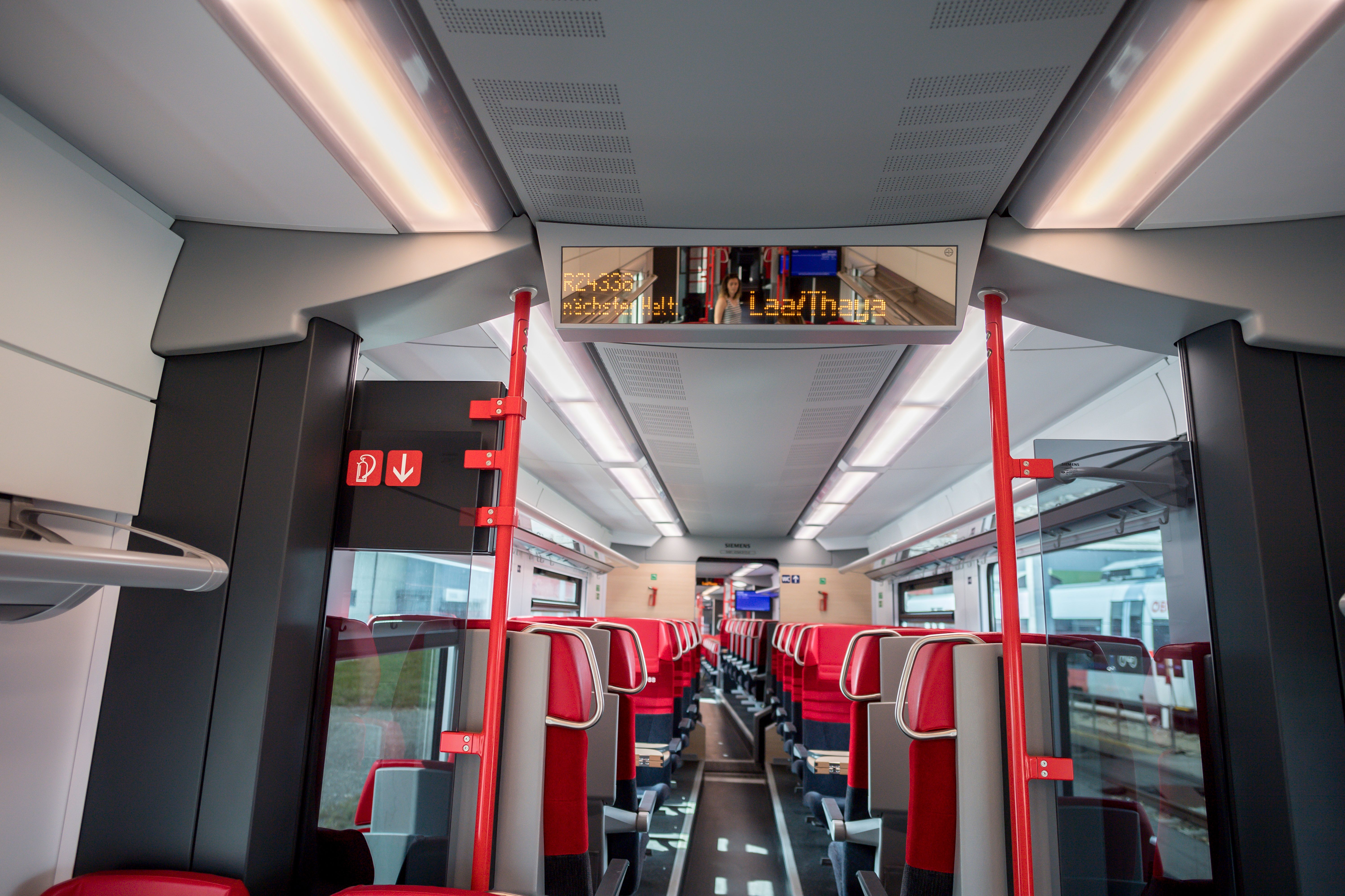 Interior view of a modern train compartment with red seats and digital display board.