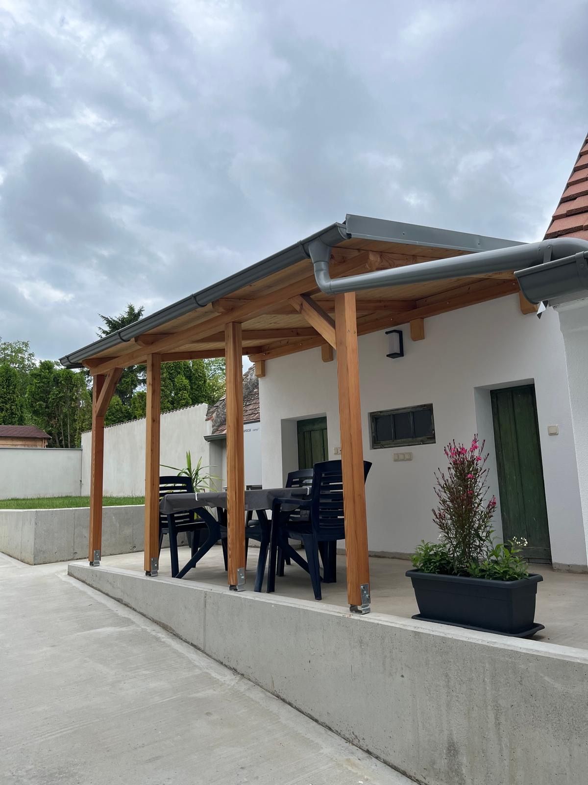 Überdachte Terrasse mit Tisch und Stühlen, umgeben von Pflanzen.